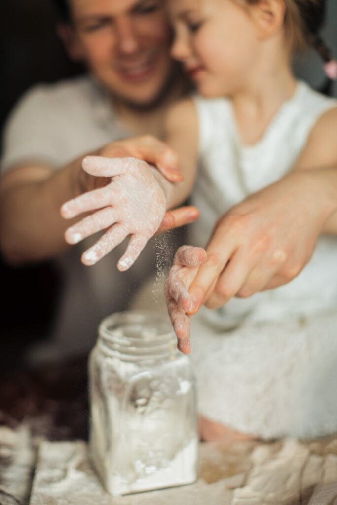 Happy crop cute girl with pigtail and blurred father shaking off flour from hands while making dough